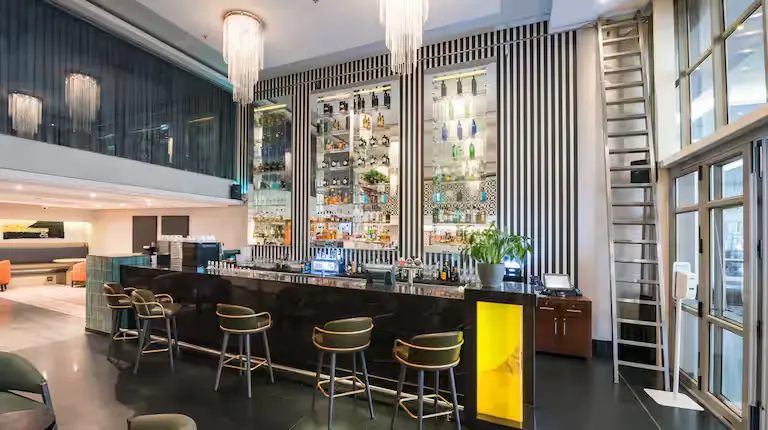 Modern hotel bar with high shelves of liquor, green chairs, a marble counter, and large hanging chandeliers in a stylish setting.