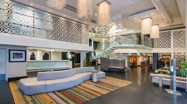 Modern hotel lobby with stylish seating, colourful striped rug, decorative wall panels, and large chandeliers, featuring a glass staircase.
