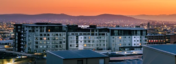 Cityscape at sunset with modern apartment blocks in the foreground and hills silhouetted against an orange sky in the background.