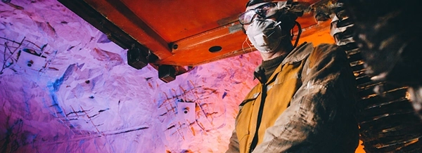 A worker in protective gear and a mask stands underground, surrounded by colourful, illuminated rock walls and metal machinery.